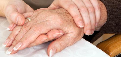 Zwei Hände, eine ältere und eine jüngere, liegen auf einem Tisch, wobei die jüngere Hand die ältere sanft umschließt.